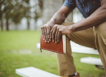 A closeup shot of a male holding the bible while sitting on a park table with a blurred background