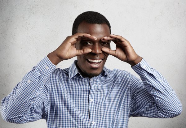 Studio portrait of handsome dark-skinned employee wearing checkered shirt holding his hands at his eyes as if looking through binoculars or glasses, smiling happily against studio wall background